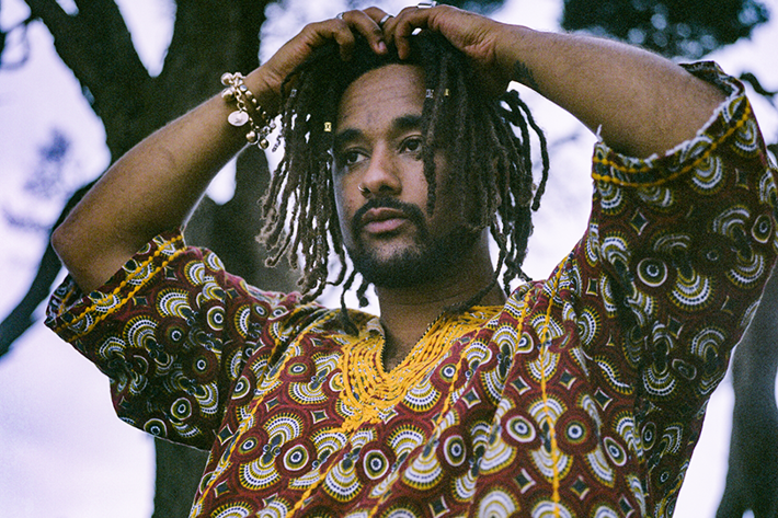 A person with dreadlocks adjusts their hair, wearing a colourful patterned top, standing outdoors among trees.