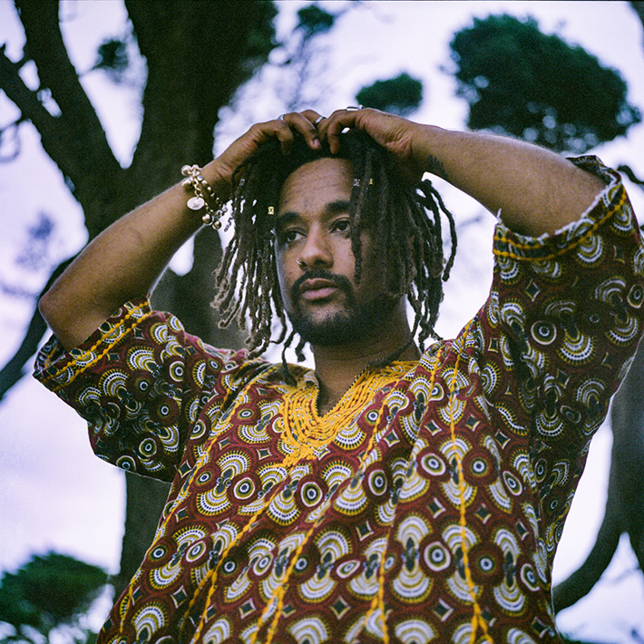 A person with dreadlocks adjusts their hair, wearing a colourful patterned top, standing outdoors among trees.