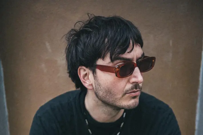 By Ian Laidlaw. A person is seated on a concrete step against a textured wall, wearing a black long-sleeve top and dark pants. Accessories include a beaded necklace, a bracelet, and rings. The individual has short, slightly tousled dark hair and sunglasses resting on the head. The overall setting has a muted, urban feel with soft natural lighting and a casual, relaxed posture.