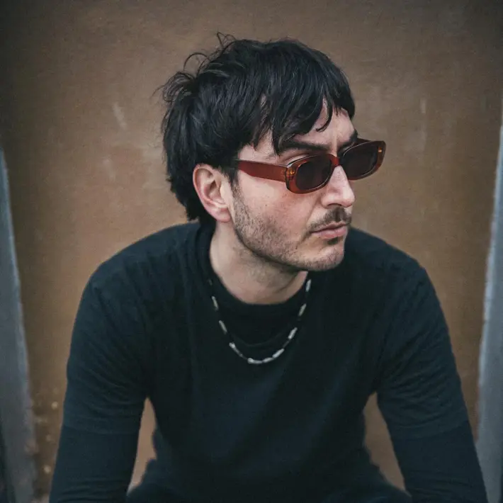 By Ian Laidlaw. A person is seated on a concrete step against a textured wall, wearing a black long-sleeve top and dark pants. Accessories include a beaded necklace, a bracelet, and rings. The individual has short, slightly tousled dark hair and sunglasses resting on the head. The overall setting has a muted, urban feel with soft natural lighting and a casual, relaxed posture.