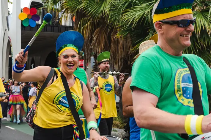 A lively street parade shows participants wearing bright yellow and green shirts with Brazilian-themed logos, playing drums and percussion instruments. They are accessorised with colourful wristbands and blue feathered headpieces. The background includes palm trees, a crowd of spectators, and a building decorated with a rainbow flower emblem, creating a festive and inclusive atmosphere.