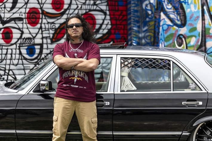 A person stands confidently with crossed arms in front of a black car, set against a colourful graffiti wall.