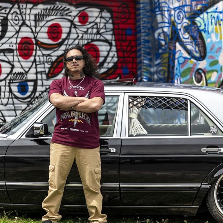A person stands confidently with crossed arms in front of a black car, set against a colourful graffiti wall.