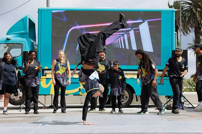 A dance crew performs an energetic street routine in front of a brightly coloured truck with a large digital screen. One dancer is balancing on one hand in a handstand while the rest of the group stands in a semicircle, watching and supporting the performance. The dancers wear coordinated black outfits with bold patterns and yellow detailing.