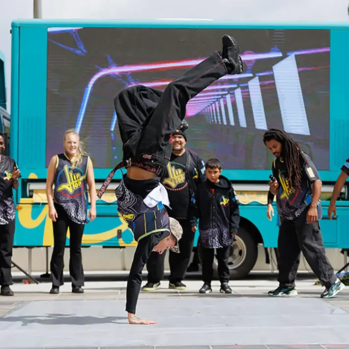 A dance crew performs an energetic street routine in front of a brightly coloured truck with a large digital screen. One dancer is balancing on one hand in a handstand while the rest of the group stands in a semicircle, watching and supporting the performance. The dancers wear coordinated black outfits with bold patterns and yellow detailing.