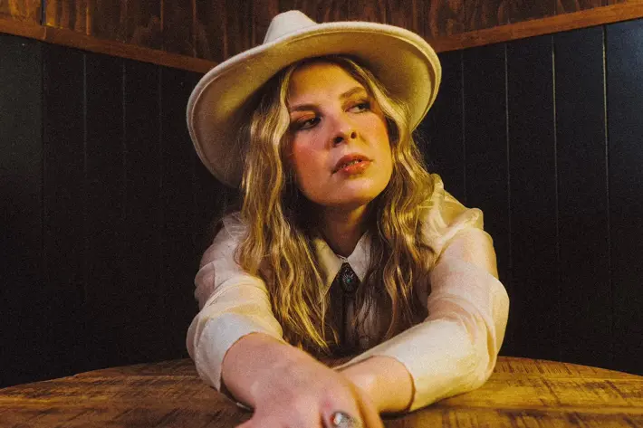 Charlie Lane sitting at table, hands out front, on table, Charlie has blonde hair and is wearing a white hat.