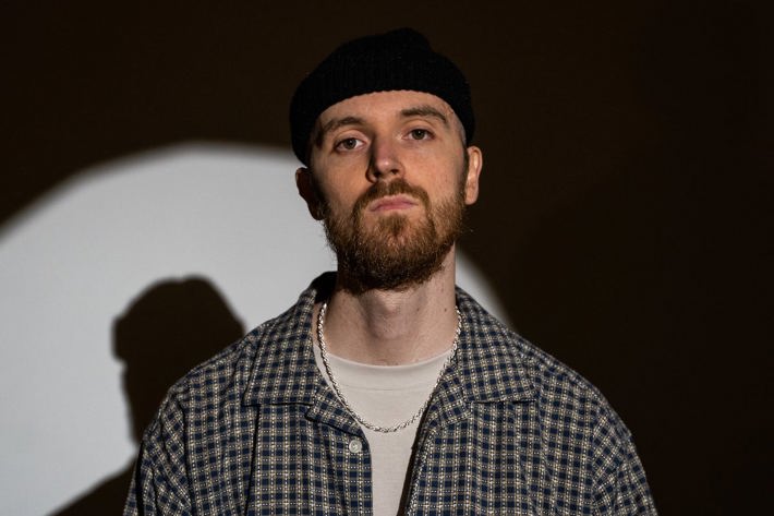 Person with a beanie and plaid shirt standing against a dark background with a circular light pattern.