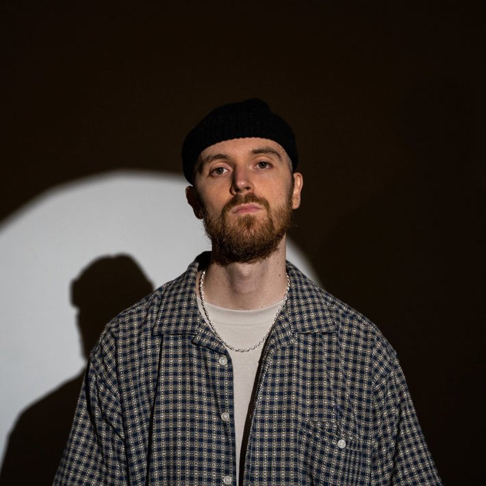 Person with a beanie and plaid shirt standing against a dark background with a circular light pattern.