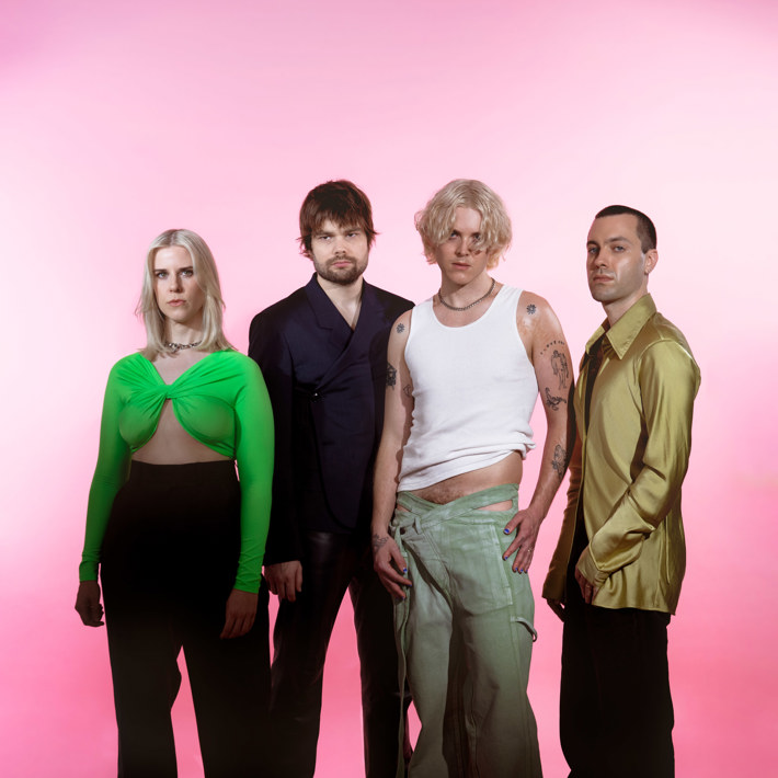 Four band members stand confidently against a soft pink background, each showcasing unique styles: a person in a bright green cut-out top, a person in a dark tailored suit, another in a white tank top and grey pants, and a fourth in a gold satin shirt.