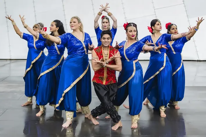A group of dancers pose outdoors in front of a modern metallic wall. They wear coordinated costumes featuring vibrant blue outfits with gold trim, while the central performer is dressed in red and black. The dancers stand barefoot on a reflective surface, with arms extended in expressive, symmetrical positions, showcasing a traditional dance style.
