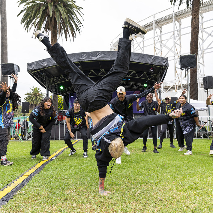 A group of dancers is performing outdoors on a grassy area in front of a stage with speakers and lighting. One dancer is in the foreground executing a handstand move, while others stand in a semicircle around, clapping and cheering. The dancers are wearing coordinated outfits with black pants and patterned tops. Tall palm trees and a large white structure are visible in the background, creating a lively and energetic atmosphere.