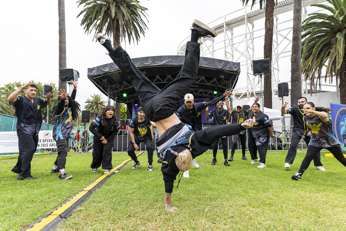 A group of dancers is performing outdoors on a grassy area in front of a stage with speakers and lighting. One dancer is in the foreground executing a handstand move, while others stand in a semicircle around, clapping and cheering. The dancers are wearing coordinated outfits with black pants and patterned tops. Tall palm trees and a large white structure are visible in the background, creating a lively and energetic atmosphere.