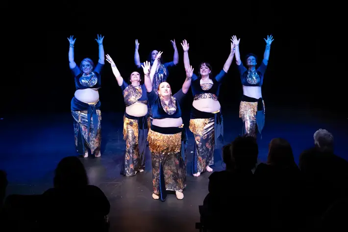 A group of performers on stage present a belly dance routine under dramatic lighting. They wear coordinated costumes featuring dark blue tops and skirts with gold accents and decorative hip scarves. The dancers stand barefoot with arms raised in expressive poses, while an audience watches from the foreground in a dimly lit theatre setting.