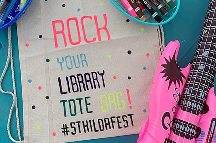 A decorated tote bag on a teal table with the text “ROCK YOUR LIBRARY TOTE BAG! #STKILDAFEST” in colourful letters and polka dots. Surrounding the bag are two baskets filled with assorted coloured markers and a bright pink inflatable guitar with black detailing.