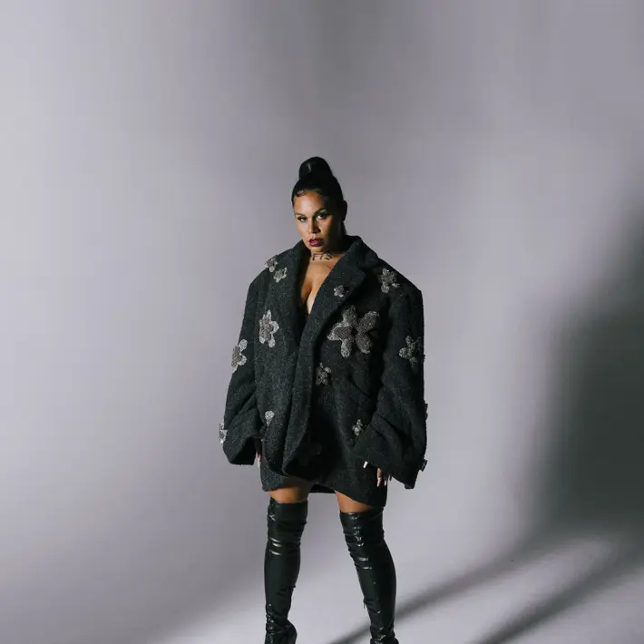 A person is standing against a plain grey backdrop wearing an oversized black textured coat decorated with large silver floral embellishments. The coat is styled as a dress, exposing the legs. The outfit is paired with thigh-high black leather stiletto boots. Soft lighting casts shadows on the floor and background, highlighting the dramatic and fashionable look.