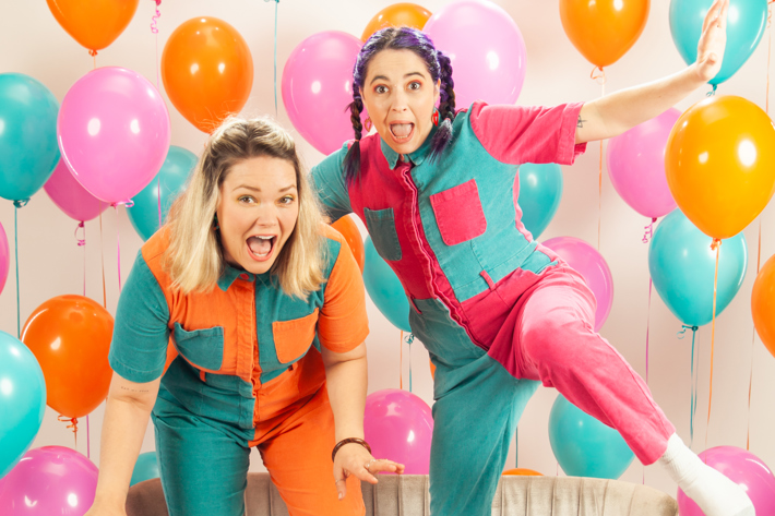 Two excited individuals in colourful jumpsuits posing on a couch surrounded by vibrant balloons.