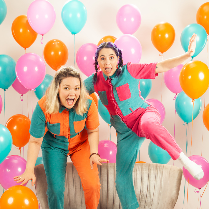 Two excited individuals in colourful jumpsuits posing on a couch surrounded by vibrant balloons.