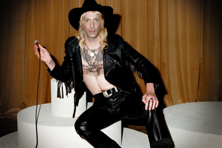 Leroy is wearing a black leather pants, open jacket fringe and black cowboy hat with thick chains around their neck.
