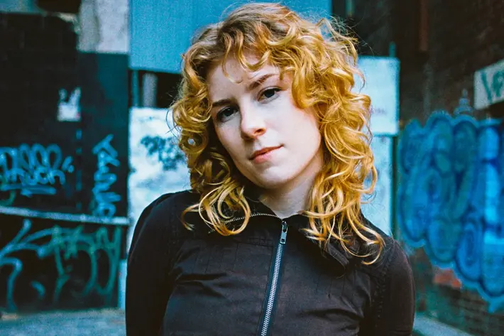 A person with curly blonde hair stands in a narrow urban alleyway with graffiti-covered walls. They are wearing a black zip-up jacket with front pockets and dark jeans, with arms positioned behind their back. The ground is paved with stone, and the background includes blue and white graffiti, a brick wall, and industrial elements like pipes and metal panels. The lighting gives the scene a gritty, street-style atmosphere.