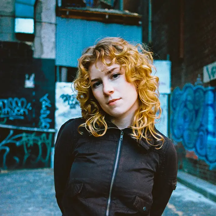 A person with curly blonde hair stands in a narrow urban alleyway with graffiti-covered walls. They are wearing a black zip-up jacket with front pockets and dark jeans, with arms positioned behind their back. The ground is paved with stone, and the background includes blue and white graffiti, a brick wall, and industrial elements like pipes and metal panels. The lighting gives the scene a gritty, street-style atmosphere.
