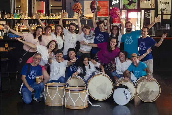 A group of people pose indoors in front of a bar, wearing colourful shirts and skirts with matching patterns. Several individuals hold large drums on the floor, while others raise their arms or hold decorative percussion instruments above their heads. The setting includes a polished dark floor, bar shelves with bottles, and vibrant wall art, creating a lively and festive atmosphere.