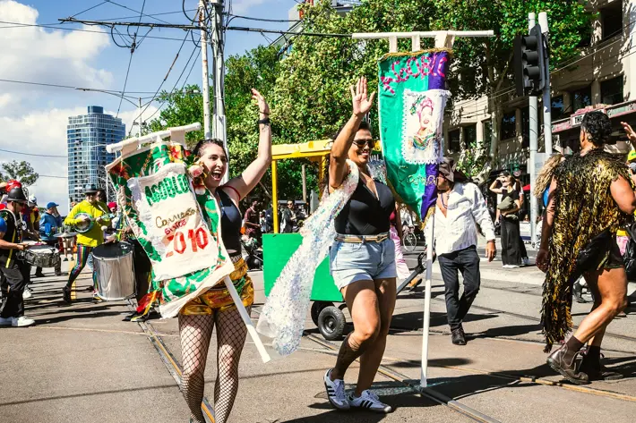 People participating in a street parade, holding colorful banners and wearing festive costumes, with others walking and playing instruments in the background.