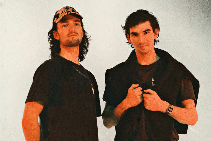 Two people are standing against a plain, light-coloured background. Both are wearing dark clothing with casual styling. One person has a patterned cap and a loose-fitting T-shirt, while the other has a short-sleeved shirt layered with a jumper draped over the shoulders and a wristwatch. The overall image has a warm, slightly grainy texture, giving it a retro or film-like aesthetic.