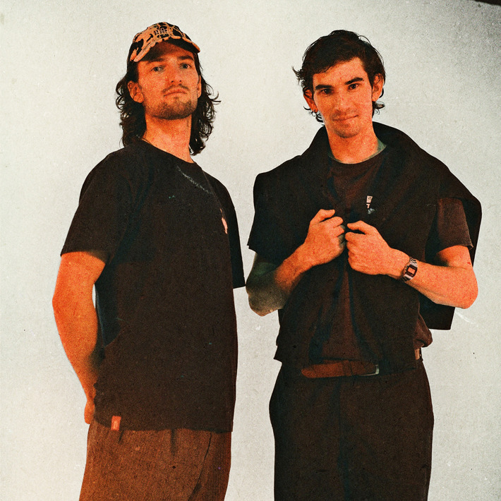 Two people are standing against a plain, light-coloured background. Both are wearing dark clothing with casual styling. One person has a patterned cap and a loose-fitting T-shirt, while the other has a short-sleeved shirt layered with a jumper draped over the shoulders and a wristwatch. The overall image has a warm, slightly grainy texture, giving it a retro or film-like aesthetic.