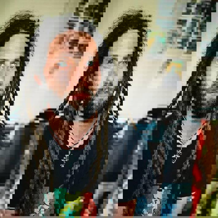 Brazaman, with long dark dreadlocks, a beard, and blue eyes. He is wearing a black t-shirt and appears to be in a room with a wall covered in posters.