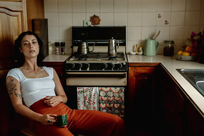 A person sits in a cosy kitchen, leaning back on a wooden chair while holding a green mug. The person wears a white fitted top and rust-coloured trousers, with a visible arm tattoo. Behind them is a gas stove with a coffee pot and kettle, patterned tea towels hanging from the oven door, and a benchtop with jars, a green jug, and a bowl of fruit. The warm lighting and wooden cabinetry create a homely, vintage feel.Photo by Tajette O'Halloran 