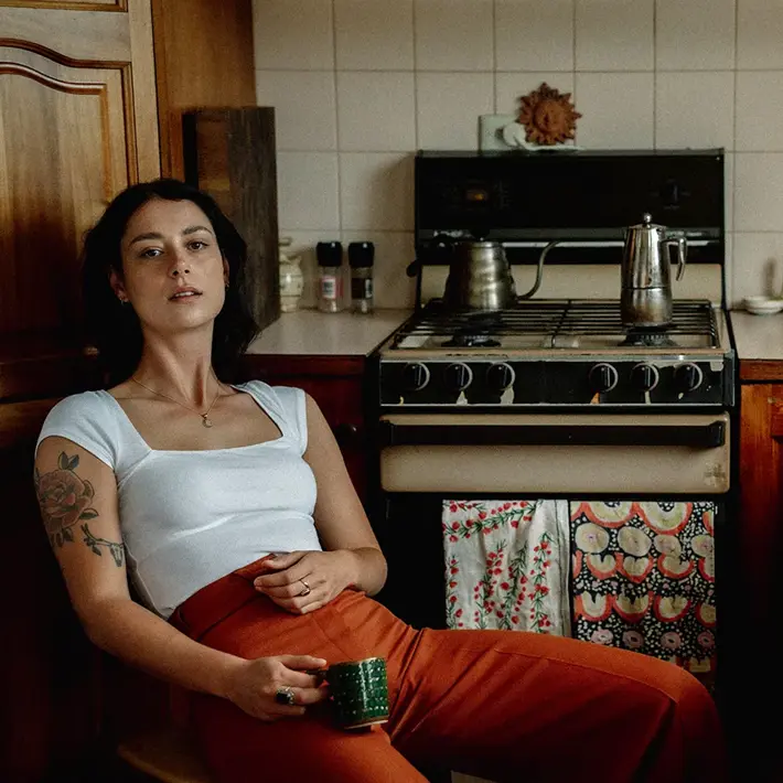 A person sits in a cosy kitchen, leaning back on a wooden chair while holding a green mug. The person wears a white fitted top and rust-coloured trousers, with a visible arm tattoo. Behind them is a gas stove with a coffee pot and kettle, patterned tea towels hanging from the oven door, and a benchtop with jars, a green jug, and a bowl of fruit. The warm lighting and wooden cabinetry create a homely, vintage feel.Photo by Tajette O'Halloran 