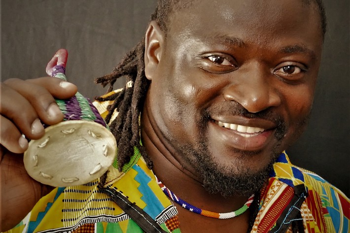 Smiling person in a colourful patterned shirt holding a traditional instrument, with a close-up focus on the face.