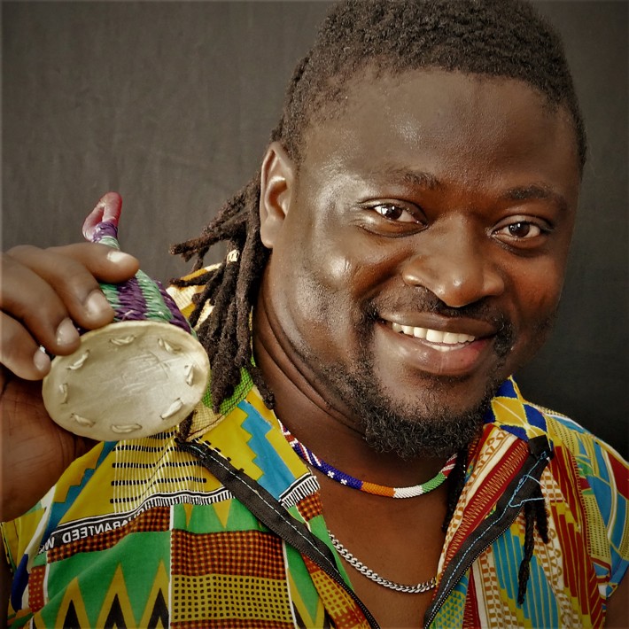 Smiling person in a colourful patterned shirt holding a traditional instrument, with a close-up focus on the face.