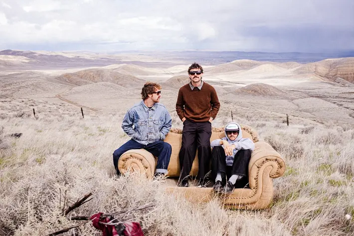 Three people sit on a tan couch placed in the middle of a dry, grassy landscape with rolling hills stretching into the distance. One person wears a denim jacket and jeans, another wears a brown jumper with dark trousers, and the third wears a light shirt with black pants and sneakers. The scene is outdoors under a cloudy sky, creating a surreal contrast between the indoor furniture and the vast, rugged terrain.
