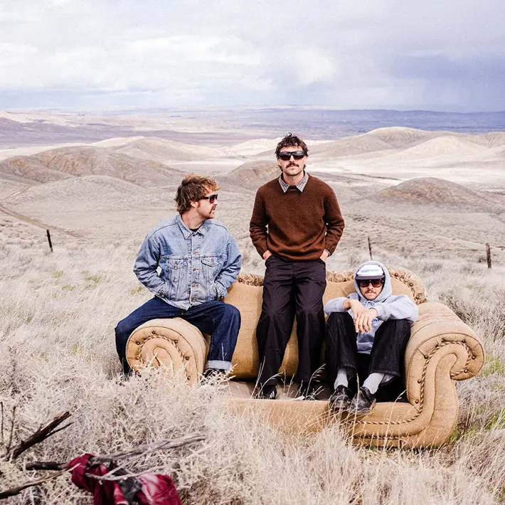 Three people sit on a tan couch placed in the middle of a dry, grassy landscape with rolling hills stretching into the distance. One person wears a denim jacket and jeans, another wears a brown jumper with dark trousers, and the third wears a light shirt with black pants and sneakers. The scene is outdoors under a cloudy sky, creating a surreal contrast between the indoor furniture and the vast, rugged terrain.