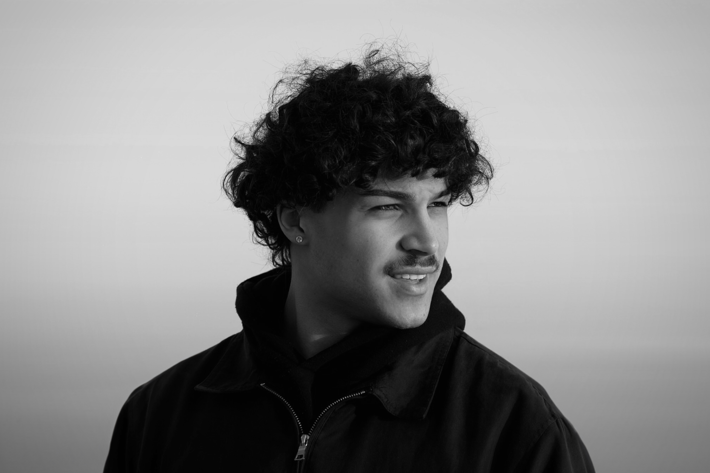 Black-and-white portrait of a person with curly hair and a moustache, looking to the side with a smile.