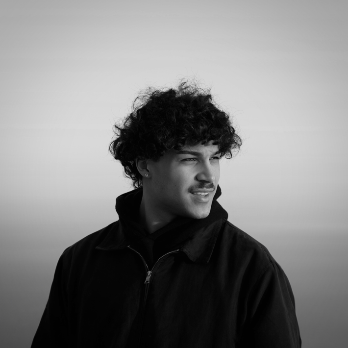 Black-and-white portrait of a person with curly hair and a moustache, looking to the side with a smile.