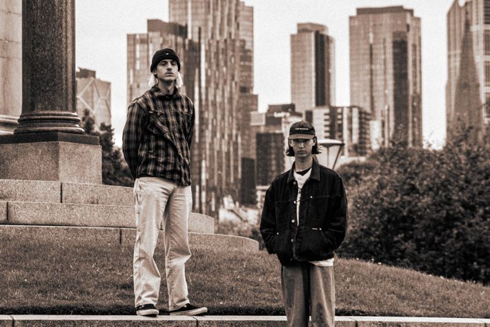 Two individuals stand on stone steps near a column, with a city skyline in the background, captured in a sepia-toned style