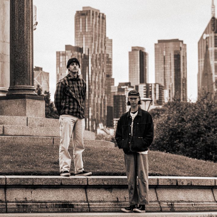 Two individuals stand on stone steps near a column, with a city skyline in the background, captured in a sepia-toned style