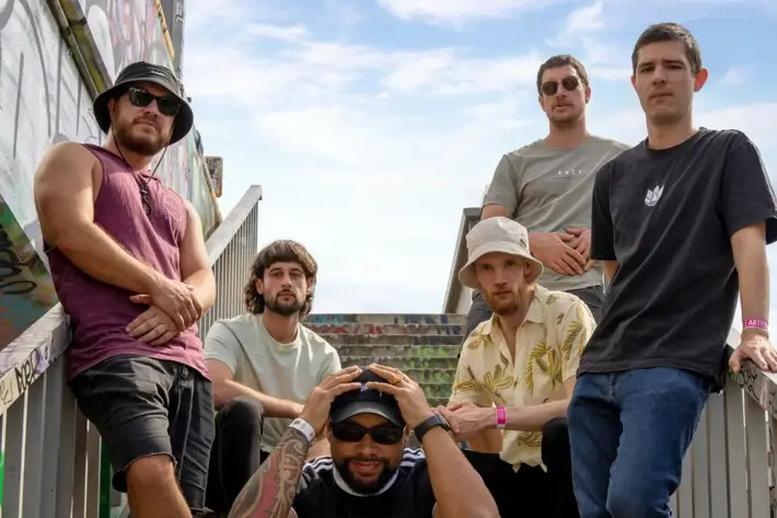 Five people pose on a graffiti-covered outdoor staircase under a partly cloudy sky. One person sits on the bottom step wearing a black T-shirt, dark shorts, striped socks, and sneakers, with tattooed arms resting on their head. The others stand or lean on the railings, dressed casually in T-shirts, jeans, and shorts, with one wearing a patterned button-up shirt and another in a sleeveless top. The setting has an urban, street-style vibe with colourful graffiti on the walls and steps.