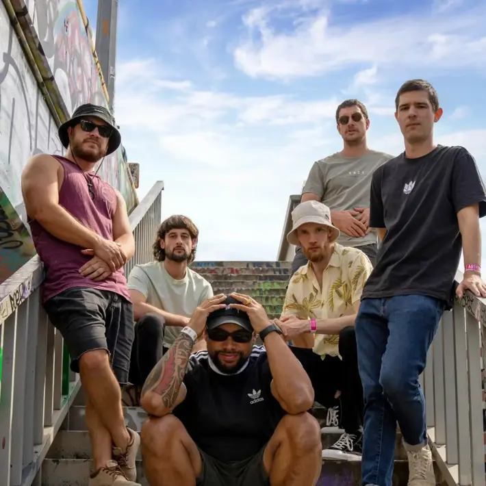 Five people pose on a graffiti-covered outdoor staircase under a partly cloudy sky. One person sits on the bottom step wearing a black T-shirt, dark shorts, striped socks, and sneakers, with tattooed arms resting on their head. The others stand or lean on the railings, dressed casually in T-shirts, jeans, and shorts, with one wearing a patterned button-up shirt and another in a sleeveless top. The setting has an urban, street-style vibe with colourful graffiti on the walls and steps.