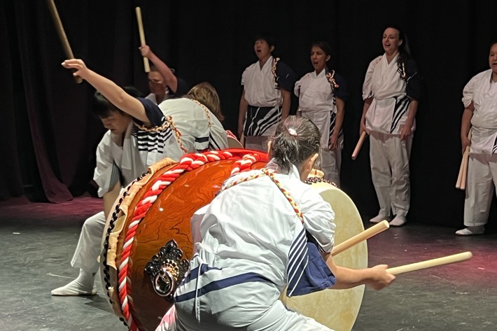 Performers in traditional Japanese attire play large drums with sticks on stage, while others sing in the background under dim lighting.