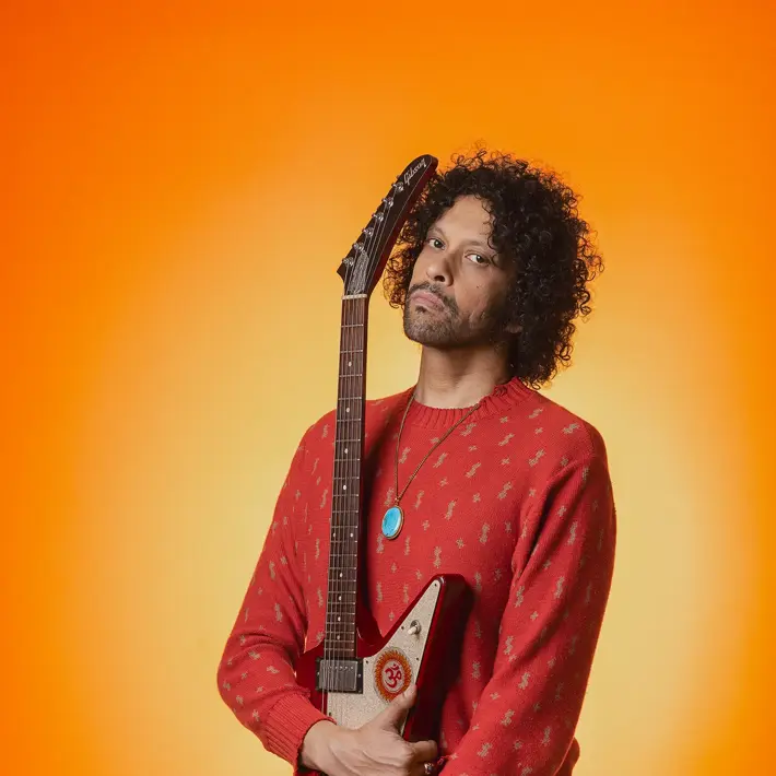 A person stands against a bright orange background, holding a red electric guitar vertically with the neck resting on the shoulder. The person is wearing a red patterned sweater, ripped black jeans, a turquoise pendant necklace, and a wristwatch. The vibrant backdrop and bold colours create a striking, high-energy portrait with a strong focus on the guitar and outfit details. Sanny Veloo is holding a 1991 Gibson Explorer for his debut album photoshoot.