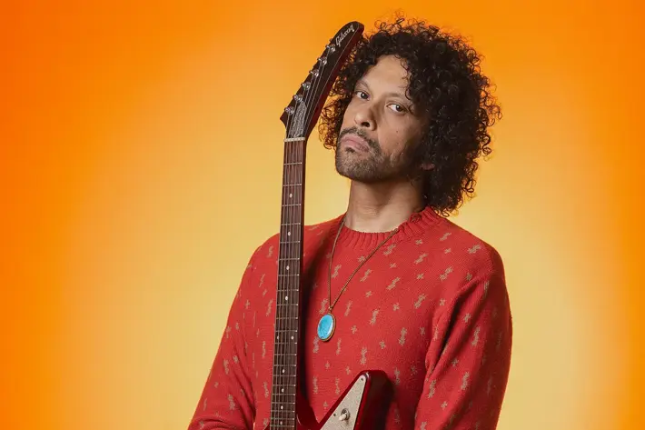 A person stands against a bright orange background, holding a red electric guitar vertically with the neck resting on the shoulder. The person is wearing a red patterned sweater, ripped black jeans, a turquoise pendant necklace, and a wristwatch. The vibrant backdrop and bold colours create a striking, high-energy portrait with a strong focus on the guitar and outfit details. Sanny Veloo is holding a 1991 Gibson Explorer for his debut album photoshoot.