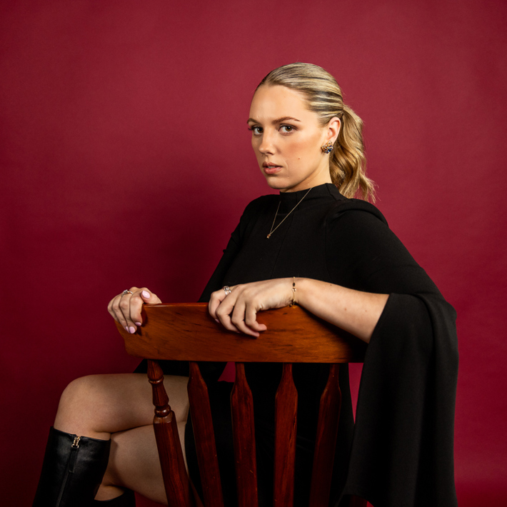 The image shows Canisha with blonde hair wearing a black outfit seated on a wooden chair against a red background. They have a serious expression on their face as they gaze directly at the camera.