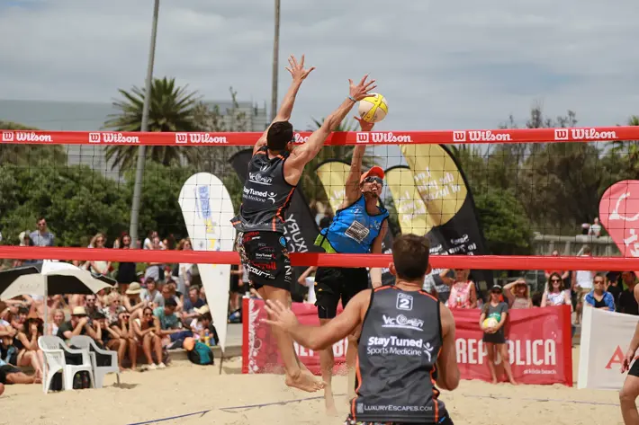 Two beach volleyball players jump at the net, reaching for the ball during a competitive match on sand. One player wears a blue top and cap, while the other wears a dark sleeveless uniform. A third player stands nearby, watching the play. The court is surrounded by banners, spectators seated on chairs, and palm trees in the background under a partly cloudy sky.