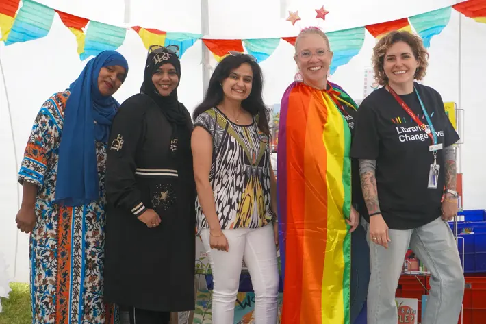 Five people stand together inside a decorated marquee with colourful bunting and star decorations. One person is wearing a rainbow pride cape, and another is wearing a black shirt with text that reads “Libraries Change Lives.” Others are dressed in patterned and traditional clothing. Brightly coloured boxes and books are visible in the background.