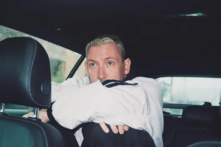 A person is sitting in the back seat of a car on a rainy day, with water droplets visible on the window. They are wearing a light-coloured long-sleeve shirt and have their arms resting on their knees, which are drawn up towards their chest. The interior of the car is dark, with black leather seats and a headrest in the foreground. The overall scene has a moody, reflective atmosphere with soft natural light coming through the windows.