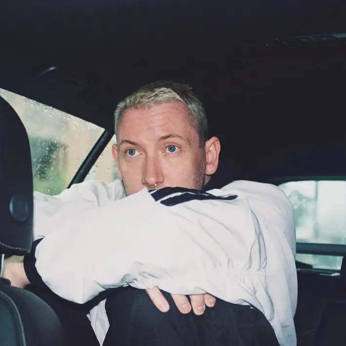 A person is sitting in the back seat of a car on a rainy day, with water droplets visible on the window. They are wearing a light-coloured long-sleeve shirt and have their arms resting on their knees, which are drawn up towards their chest. The interior of the car is dark, with black leather seats and a headrest in the foreground. The overall scene has a moody, reflective atmosphere with soft natural light coming through the windows.