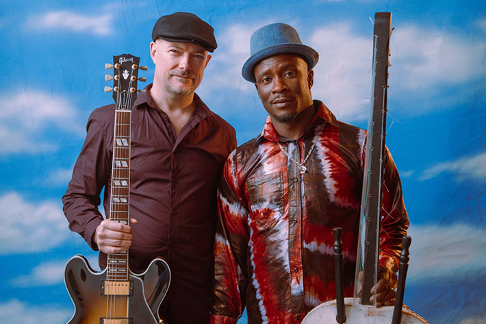 Two men wearing hats stand facing the camera, in front of a backdrop with blue skies and clouds. The men are holding a guitar and a kora (African harp). 